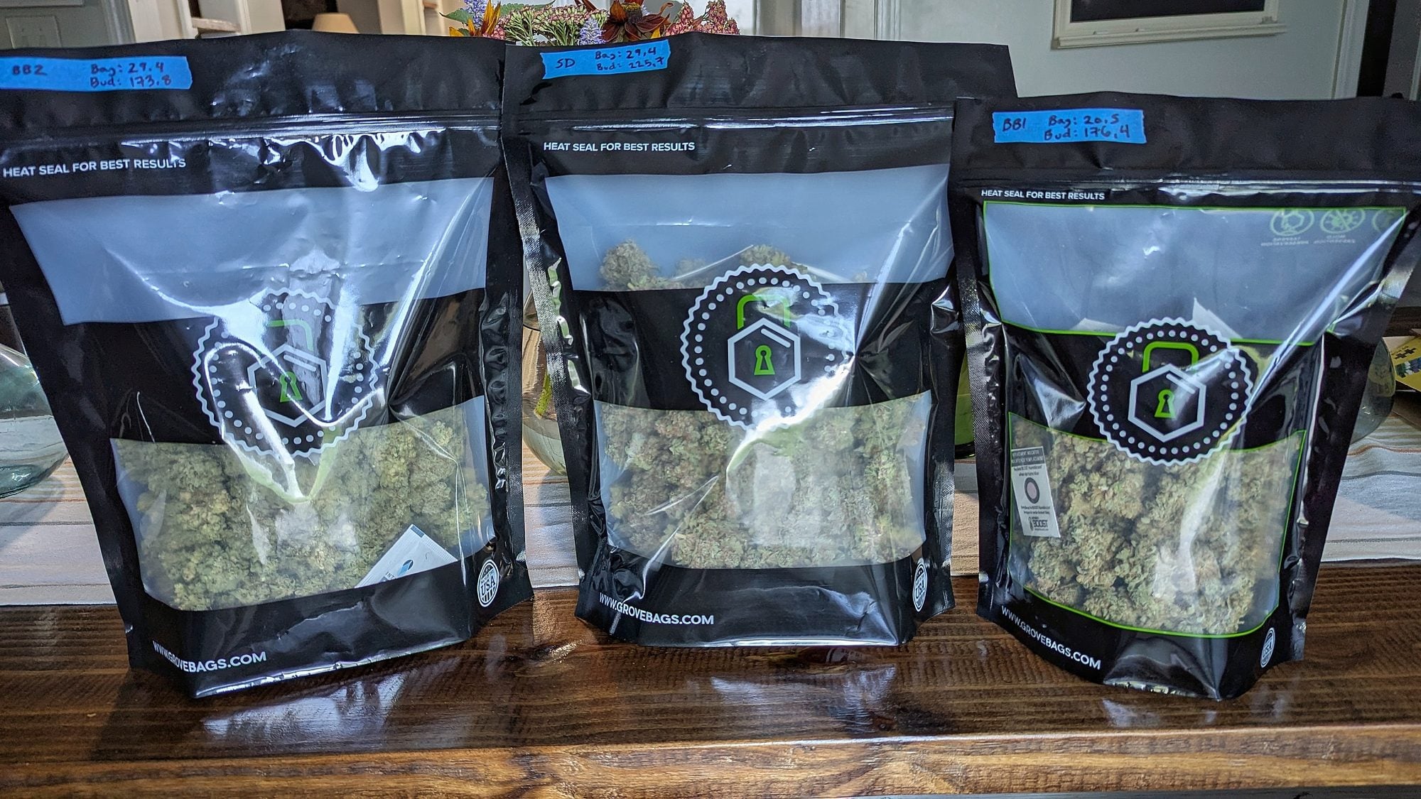 Three sealed black and white bags with hexagon logos containing dried cannabis flower with blue labels on wooden surface