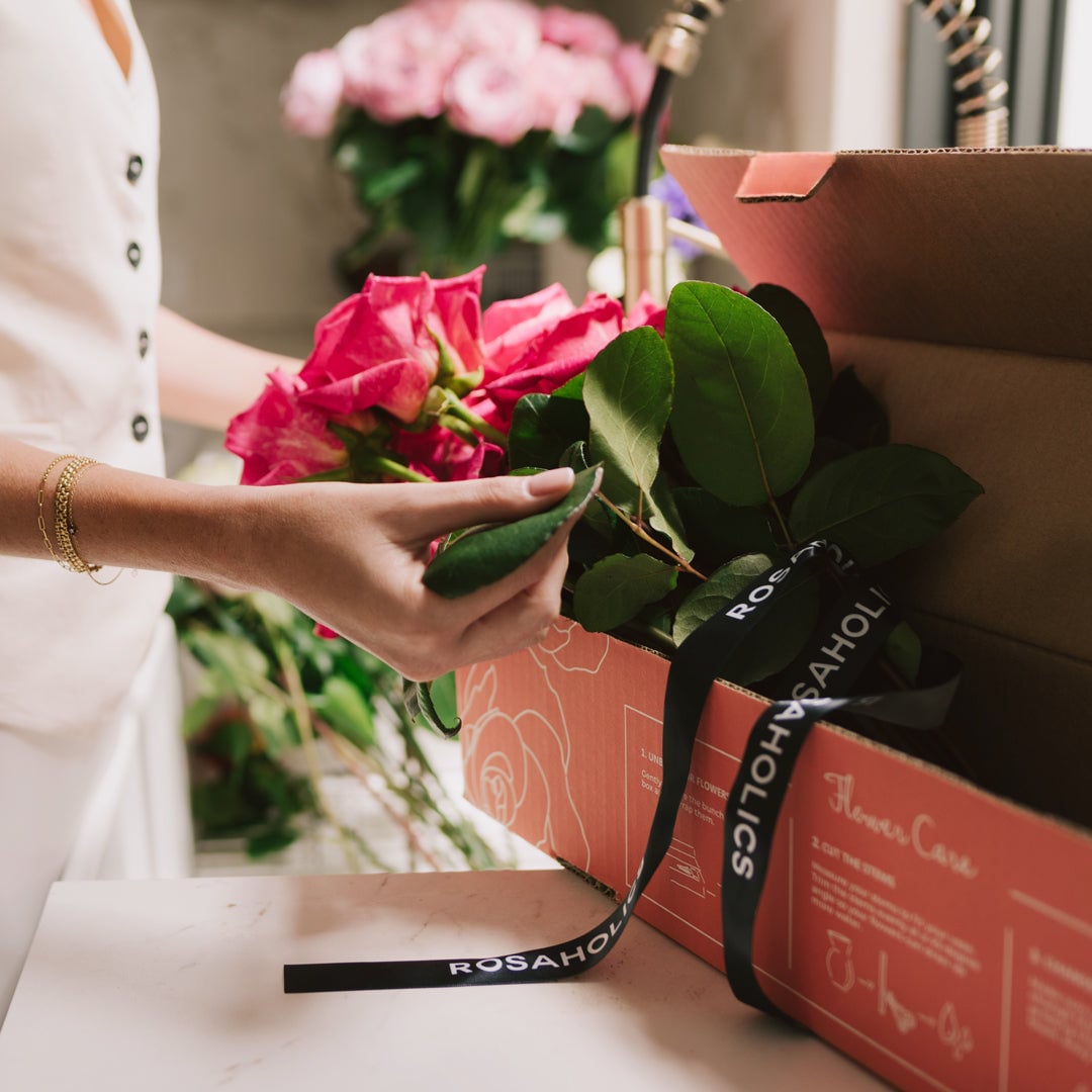 Woman in white shirt placing fresh magenta roses from a coral-colored box with black ribbon