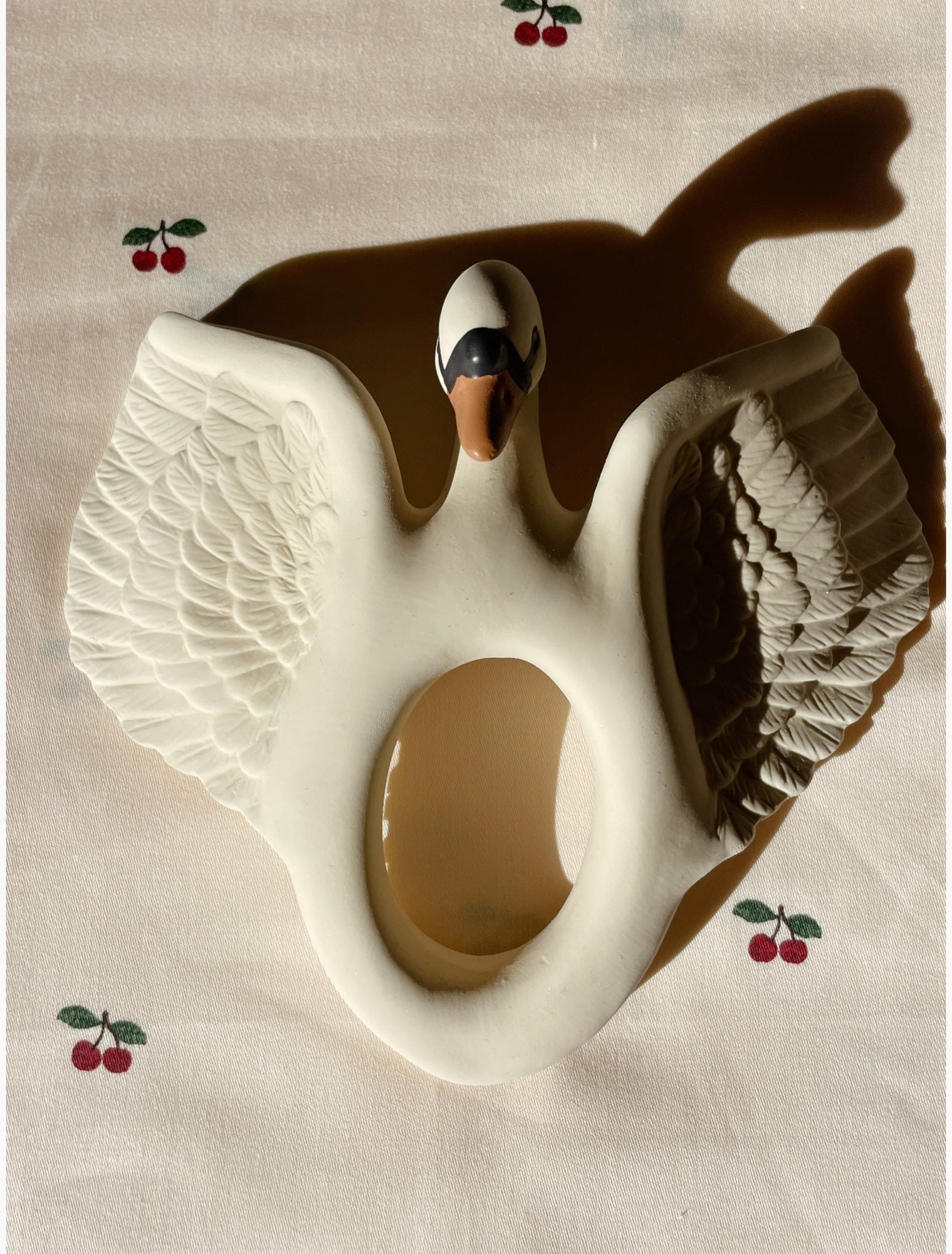 Cream ceramic swan teether with detailed feather texture, black and white head markings, and brown wings, displayed on beige fabric with embroidered cherries