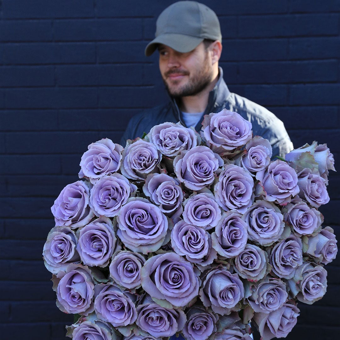 Man in gray cap and jacket holding large bouquet of lavender roses against dark brick wall