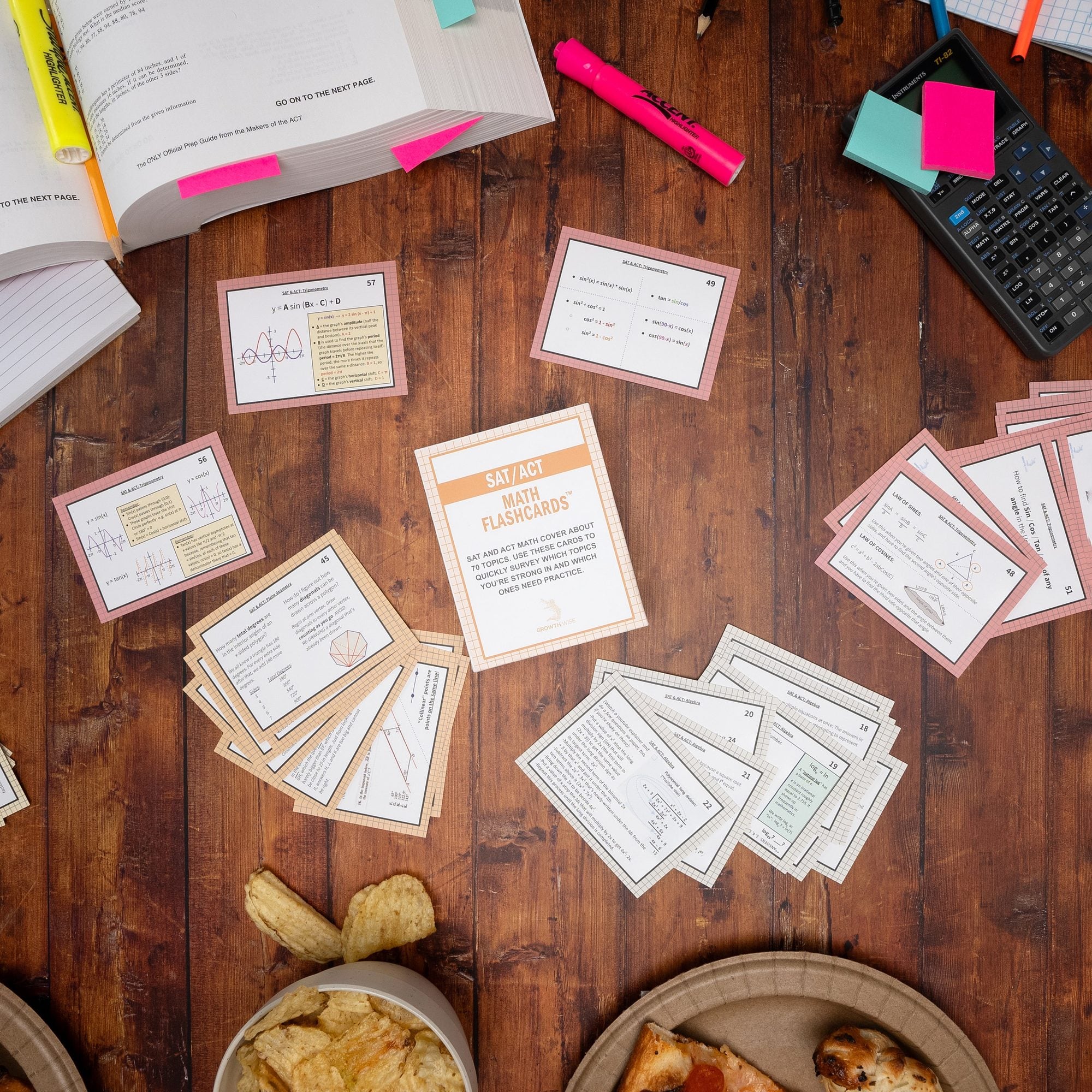 Overhead view of SAT and ACT math flashcards with orange borders spread across wooden table alongside pink highlighters, notebooks, and snack bowls
