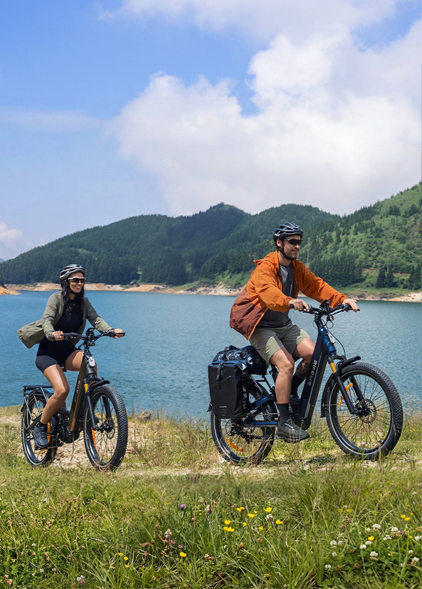 Two cyclists riding electric bikes along a grassy lakeside path with forested mountains in the background