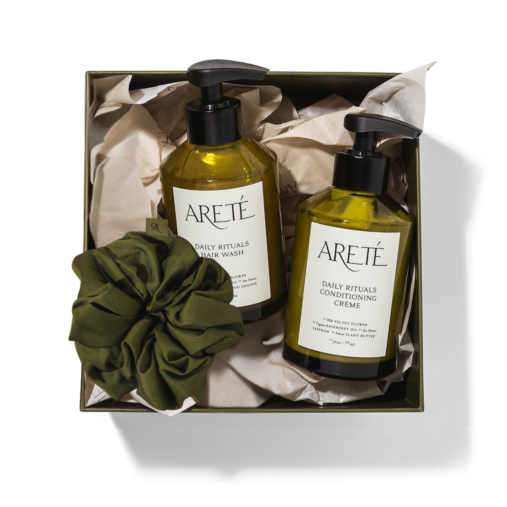 Olive-toned gift box containing two green-labeled hair care bottles with pump dispensers, cream-colored tissue paper, and an olive silk scrunchie