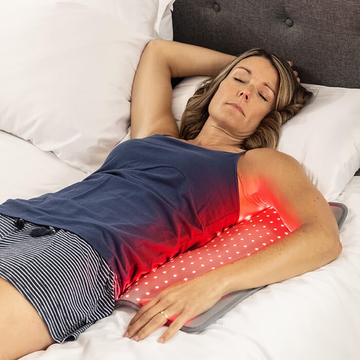 Woman lying in bed with hand on hip, red light therapy pad glowing on lower body
