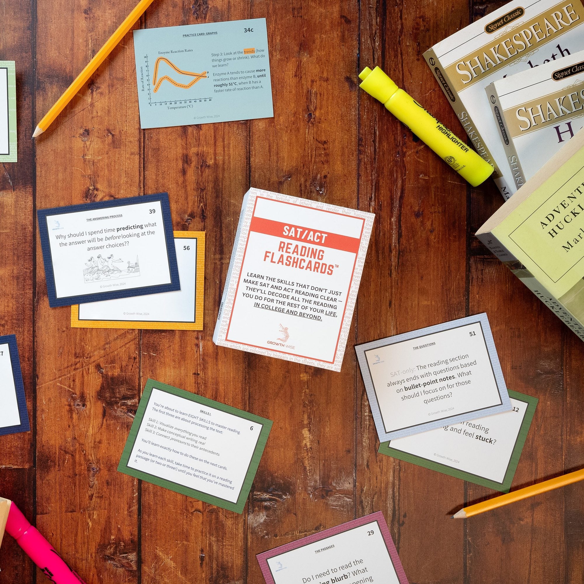 Scattered study materials on wooden desk including SAT reading flashcards, practice problem cards, highlighters, pencils, Shakespeare books, and a calculator